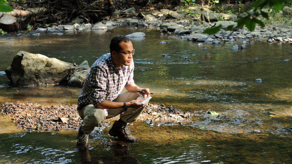 Student crouches at the edge of a river to test water quality