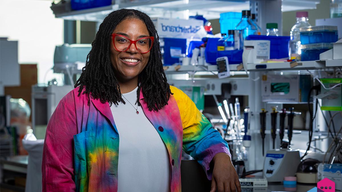 Jasmine King wearing a white shirt and rainbow-colored blazer in front of research equipment.